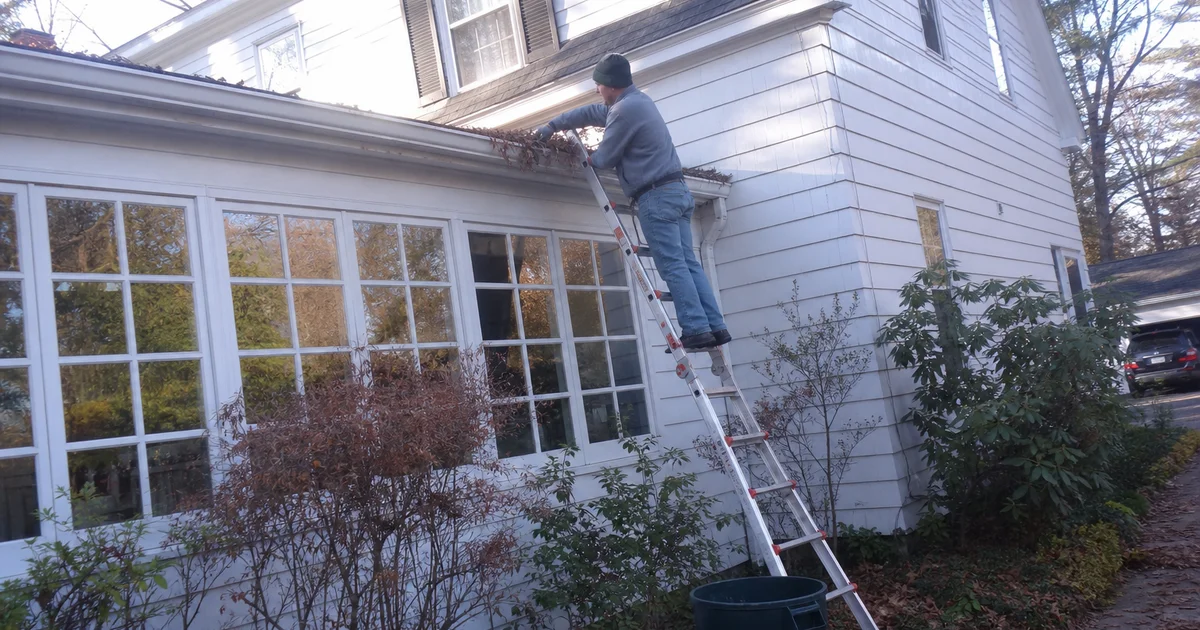 Handyman clearing wet leaves from a colonial home's gutters in West Hartford during a fall afternoon